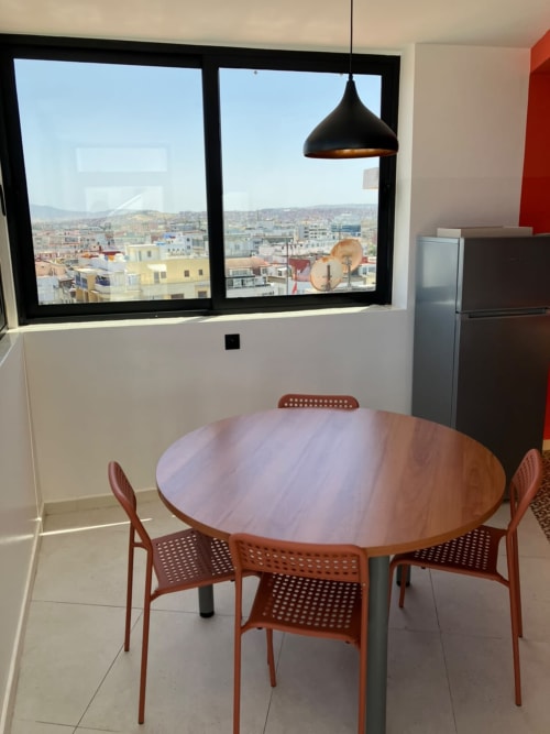 Modern dining area with wooden round table and red chairs overlooking Tangier cityscape through large black-framed windows at