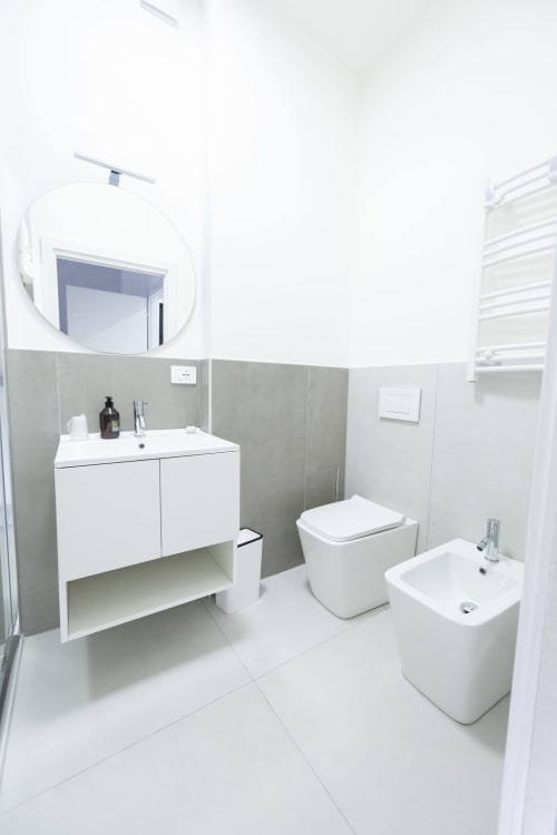 Bagno moderno dell'hotel Newhouse a Bari con sanitari bianchi, rivestimento in piastrelle grigie, specchio rotondo e scaldasalviette