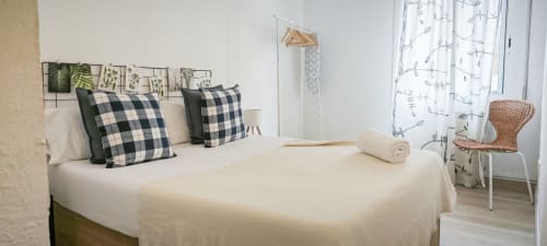 Moderno dormitorio en BCNMADflats Barcelona con almohadas de cuadros, ropa blanca, arte de pared vegetal y silla de rattán