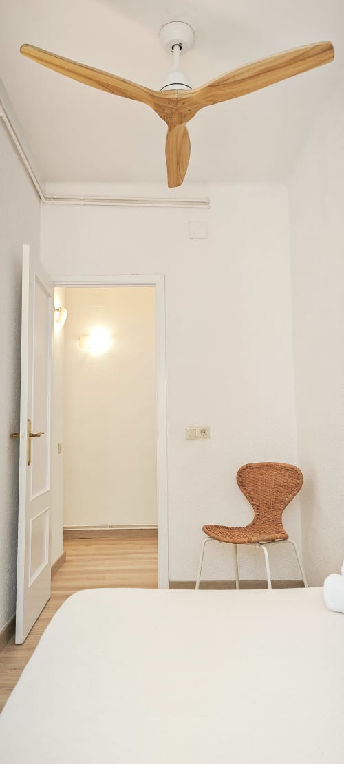 Dormitorio moderno en apartamento barcelonés con ventilador de techo de madera, paredes blancas, silla de mimbre y puerta abierta a pasillo luminoso