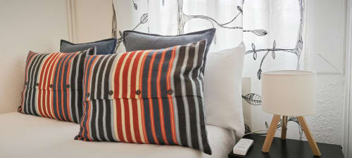 Almohadas naranja y gris a rayas en cama blanca del apartamento BCNMADflats Barcelona con cortinas botánicas y lámpara moderna