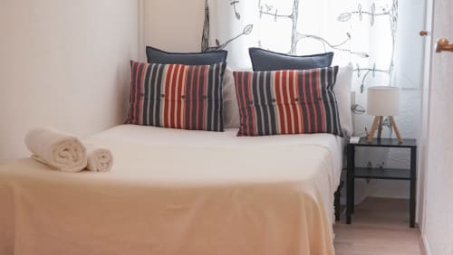 Dormitorio moderno de apartamento barcelonés con almohadas de rayas rojo y gris, ropa de cama blanca, toallas enrolladas y mesita de noche negra