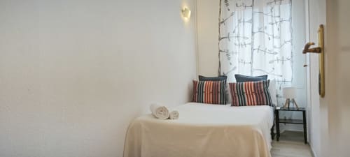 Luminoso dormitorio de apartamento barcelonés con ropa de cama blanca, almohadas rayadas, cortinas modernas y mesilla compacta