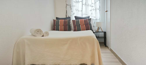 Dormitorio moderno en apartamento de Barcelona con almohadas rayadas, ropa de cama blanca, toallas enrolladas y luz natural de la ventana