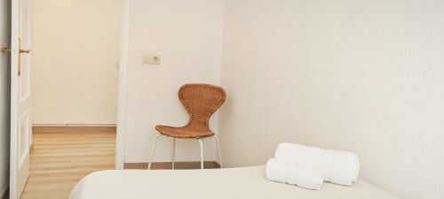 Dormitorio minimalista en apartamento de Barcelona con silla tejida, cama, almohadas blancas y piso de madera