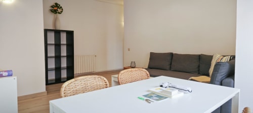 Moderno salón de apartamento en Barcelona con mesa de comedor blanca, sillas tejidas, sofá oscuro, estantería negra y suelos de madera