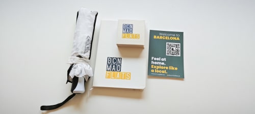 Kit de bienvenida BCNMADflats con paraguas, guías de apartamentos y tarjeta QR sobre fondo blanco