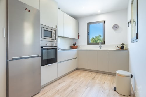 Cocina moderna con muebles blancos, electrodomésticos de acero inoxidable, suelos de madera y ventana con vistas al jardín en villa de Artà
