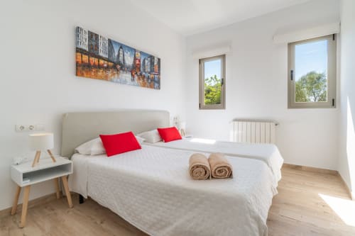 Dormitorio moderno y luminoso con cama blanca, almohadas rojas y toallas enrolladas en la villa Can Colomer Ferragut de Artà