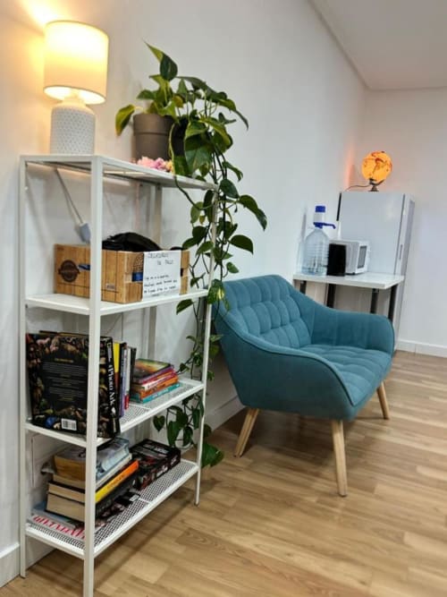 Acogedor rincón de lectura en Hostal Zaragoza Centro con sillón turquesa, estantería blanca, planta en maceta e iluminación cálida