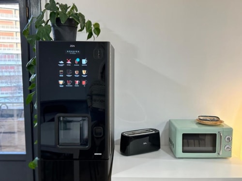 Sala de descanso del Hostal Zaragoza Centro con máquina expendedora de bebidas negra, tostadora, microondas menta, planta en maceta y vistas a la ciudad.