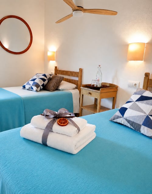 Habitació moderna a l'hotel Mas Ribas de Palamós amb llits bessons turquesa, mobles de fusta i tovalloles taronges