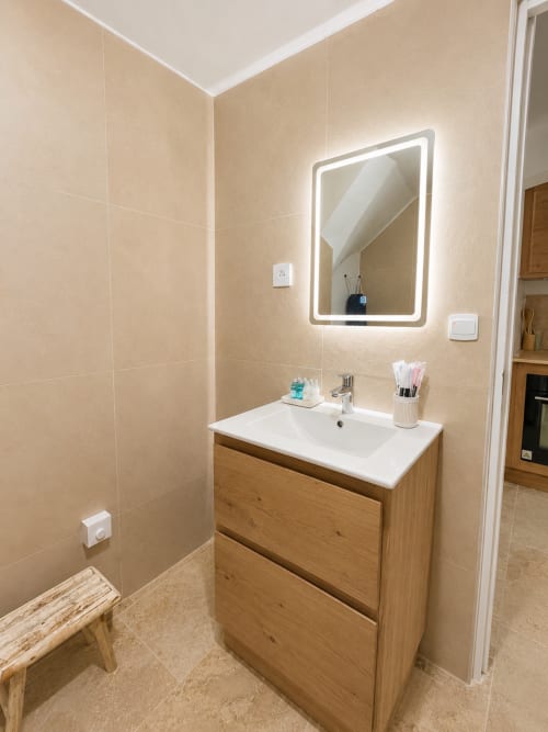Salle de bain moderne avec meuble vasque en bois, lavabo blanc, miroir lumineux et murs carrelés beige de l'appartement By Elodie, Hyères
