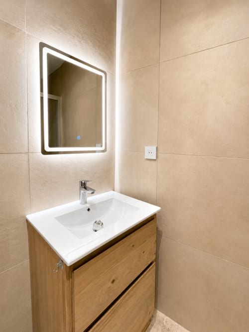 Salle de bain moderne avec miroir lumineux, lavabo blanc, vanité en bois et carrelage beige à l'appartement By Elodie à Hyères