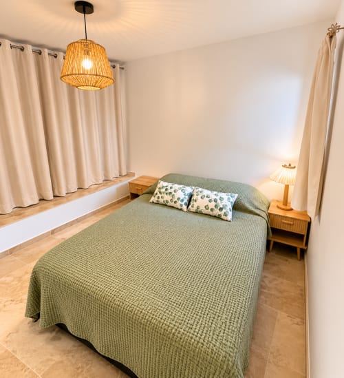 Chambre moderne avec lit en vert sauge, tables de nuit en bois, suspension tressée, rideaux crème et parquet en bois