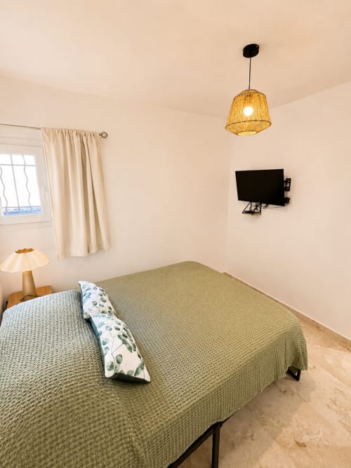 Chambre lumineuse du By Elodie à Hyères avec lit vert, lampe suspendue, TV murale et fenêtre aux rideaux beige