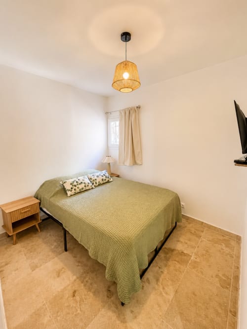 Chambre lumineuse de l'appartement By Elodie à Hyères avec literie verte, table de nuit en bois, luminaire suspendu et carrelage