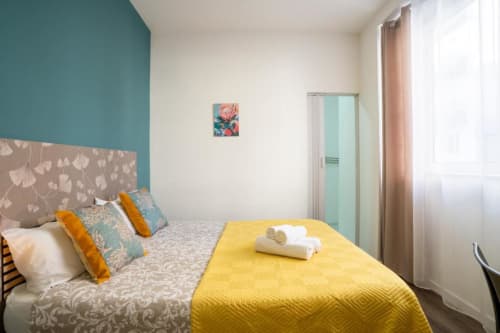 Quarto duplo em Madeirarooms Funchal com parede de destaque azul-piscina, manta amarela, almofadas coloridas e comodidades modernas