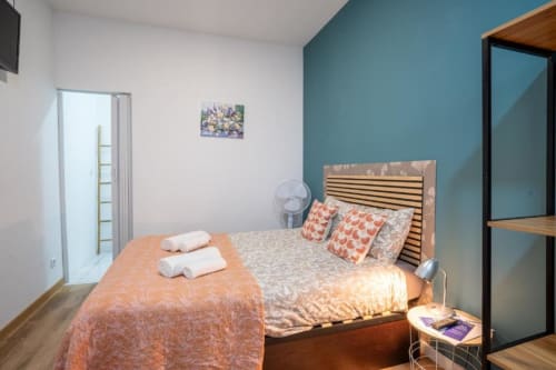 Quarto duplo em Madeirarooms Funchal com parede de destaque azul-esverdeado, cama de madeira, roupa de cama laranja e entrada do banheiro privativo.