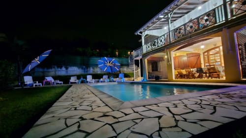 Villa de luxe avec piscine illuminée la nuit, chaises longues, parasols bleus, et maison principale éclairée avec terrasse
