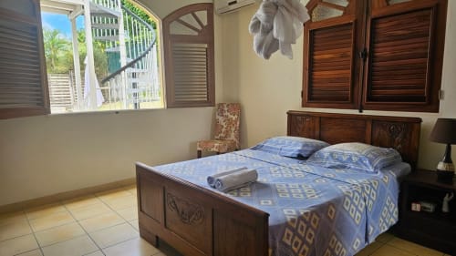Chambre spacieuse avec lit en bois, literie bleue motifs, carrelage et fenêtre donnant sur la piscine à Villa La Suite