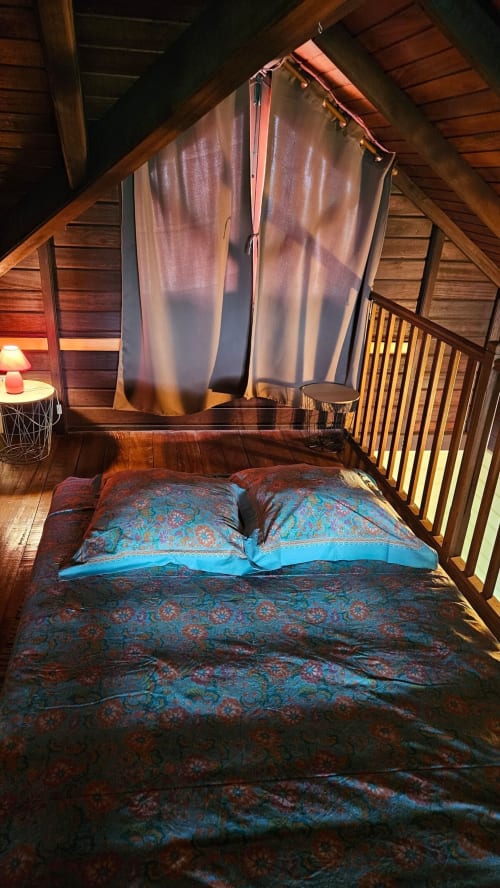 Chambre de villa en bois avec literie bleue à motifs, oreillers et voilages aérés dans une location tropicale en Martinique