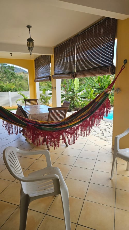 Véranda tropicale avec hamac coloré, chaises en bois et stores en bambou surplombant piscine et jardins