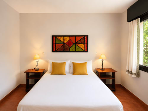 Habitación luminosa en la casa de huéspedes Vila Santa Rita, Oia, con cama blanca, almohadas doradas, mesitas de noche de madera y decoración geométrica colorida