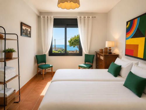 Dormitorio luminoso en la cabaña Holibai Vila Santa Rita en Oia con camas gemelas, sillas verdes, ventana con vistas al mar y arte colorido