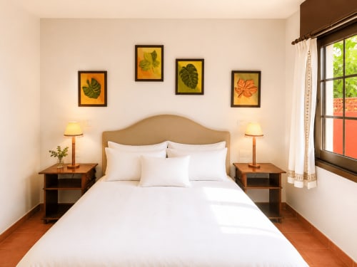 Dormitorio luminoso en la casa Holibai, Oia, con ropa de cama blanca, cabecero beige, lámparas gemelas y arte botánico en la pared