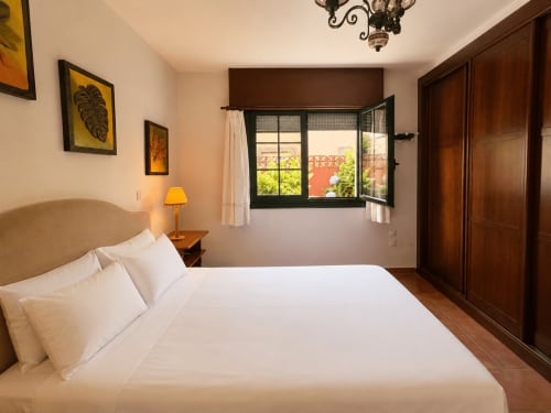 Dormitorio luminoso en cabaña Oia con ropa de cama blanca, armario de madera, ventana con vista al mar y arte botánico amarillo