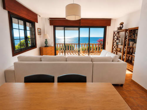 Luminoso salón con sofá seccional beige, mesa de comedor de madera y vistas espectaculares al mar de Oia a través de grandes puertas de vidrio