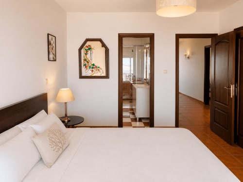 Dormitorio moderno en la casa de huéspedes Vila Santa Rita, Oia, con ropa de cama blanca, muebles de madera oscura y acceso a vistas del mar a través de cristal