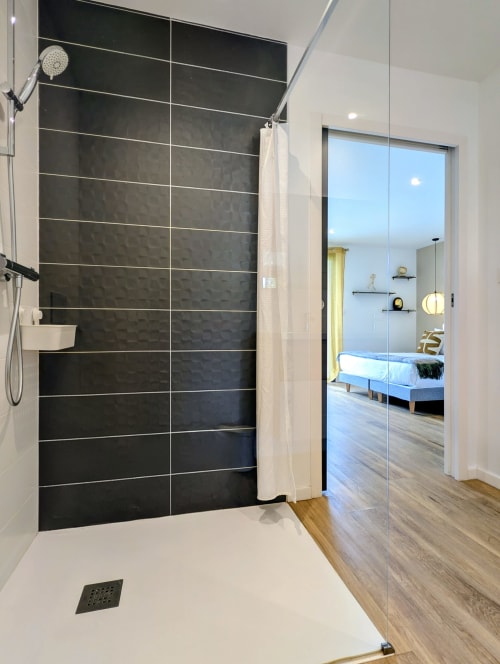 Salle de bain moderne avec douche à l'italienne carrelée de noir, paroi vitrée et vue sur la chambre au gîte La Clairière aux Chênes