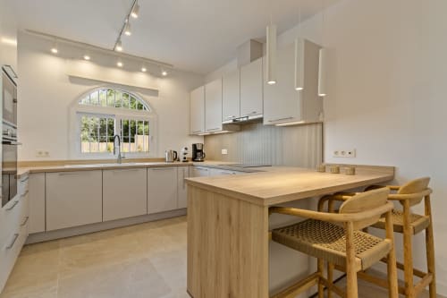 Cocina moderna con alacenas blancas, isla de madera con asientos y ventana arqueada con vistas a las montañas en Ayma Villa