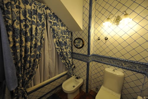 Cuarto de baño del Palacio Vasco con cortina de patrón azul, inodoro y bidé blancos, espejo redondo pequeño y paredes de azulejos