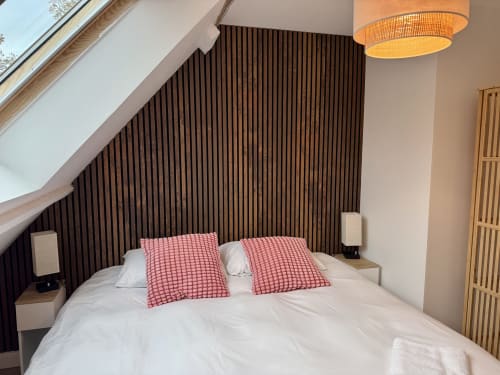 Chambre moderne avec tête de lit en lattes de bois foncé, literie blanche, coussins rouges, luminaire suspendu et plafond mansardé à la Maison Inès