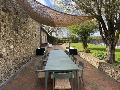 Terrasse de restaurant extérieure de la villa Maison Inès à Montigny-le-Chartif avec table longue, chaises vertes, voile d'ombrage et jardins manucurés