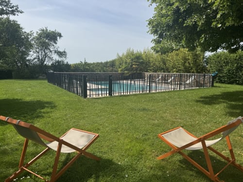 Deux chaises longues en bois face à une piscine chauffée avec clôture de sécurité noire dans le jardin verdoyant de la villa Hellofamily