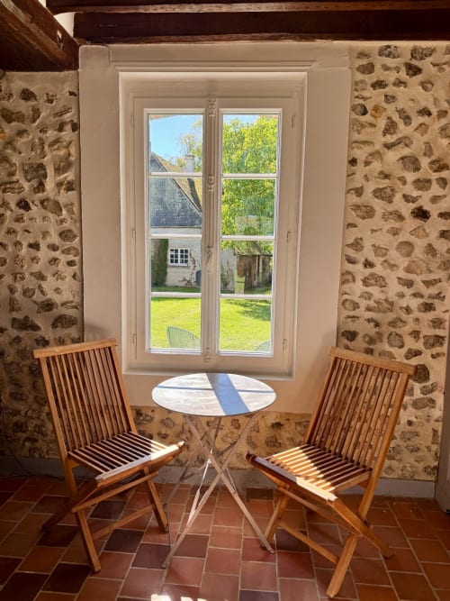 Pièce intérieure ensoleillée avec deux chaises en bois encadrant une petite table, murs en pierre, portes-fenêtres donnant sur la piscine du jardin