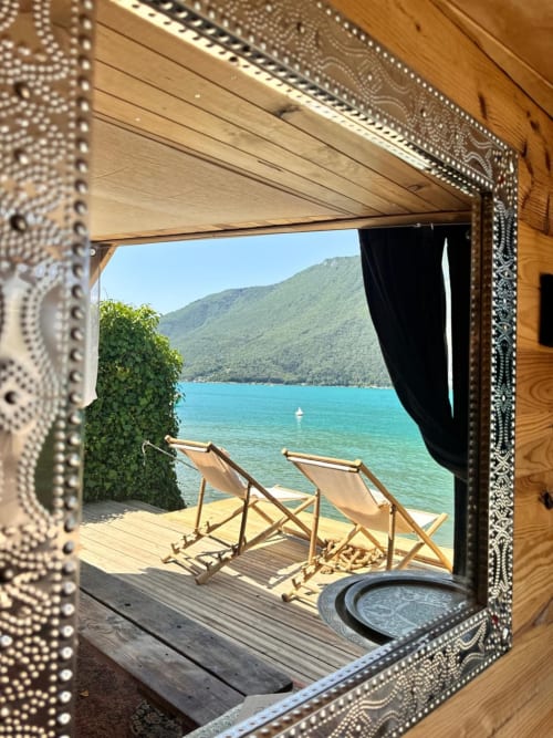 Vue encadrée du lac turquoise, transats et montagnes boisées depuis la chambre La plage secrète du gîte de Chaparon