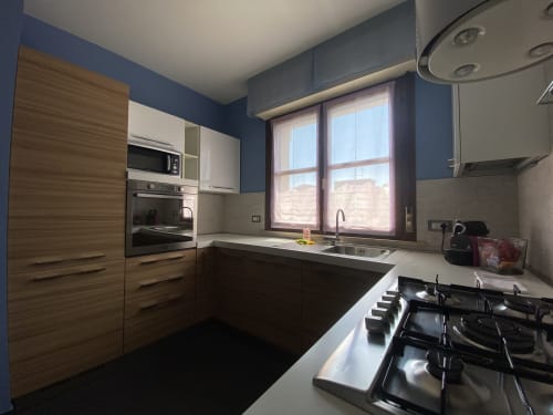 Modern kitchen with blue walls, wooden cabinetry, gas cooktop, and large windows at Cinelli Apartment in Vercelli, Italy