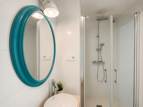 Baño moderno del hotel Holibai en Baiona con espejo turquesa, ducha tipo lluvia y accesorios blancos