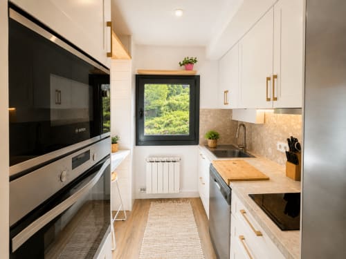 Moderna cocina blanca en Holibai con encimeras de madera, electrodomésticos de acero inoxidable y vistas a la naturaleza de Baiona