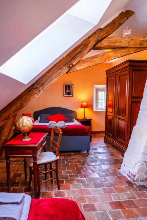 Chambre mansardée à La Maison Du Saule avec poutres apparentes, murs orange, literie rouge, mobilier en bois et lucarne