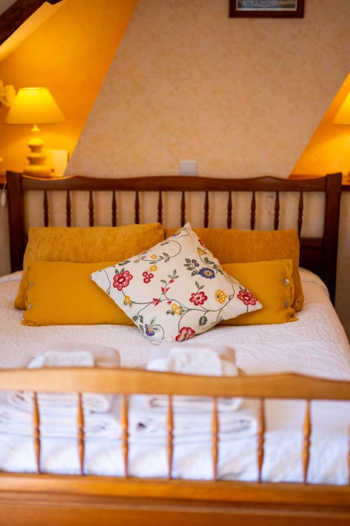 Chambre douillette à La Maison Du Saule avec lit en bois, draps blancs, oreillers dorés et coussin fleuri