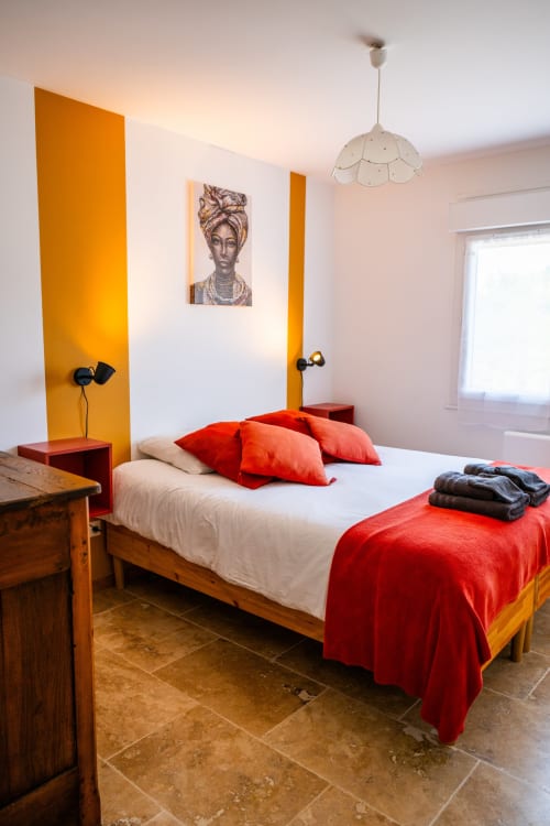 Chambre moderne de la Maison Du Saule avec accents orange vifs, lit en bois, coussins rouges et portrait abstrait
