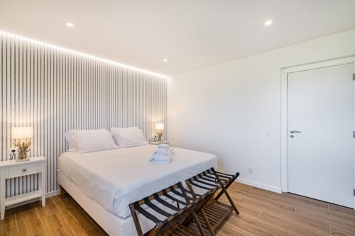 Quarto moderno e iluminado na Atlantic Places Albufeira com cama branca, cabeceira com padrão listrado, iluminação ambiente, piso de madeira e
