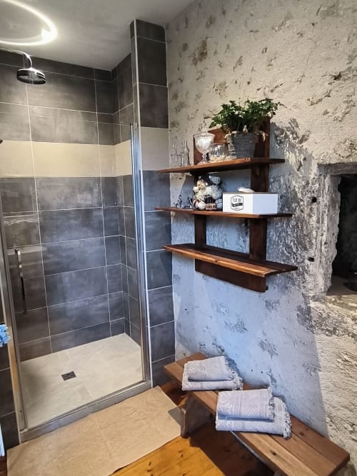 Salle de bain moderne avec douche à l'italienne, murs en pierre brute, étagères en bois et serviettes pliées aux 2 Brigards B&B