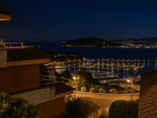 El hotel Holibai domina la marina iluminada de Baiona por la noche con embarcaciones y montañas costeras